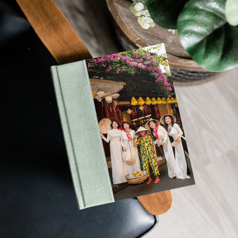 A hardcover photo album with a light sage green fabric cover and a printed photograph of women in traditional Vietnamese clothing, shown resting on a wooden chair next to a potted plant.