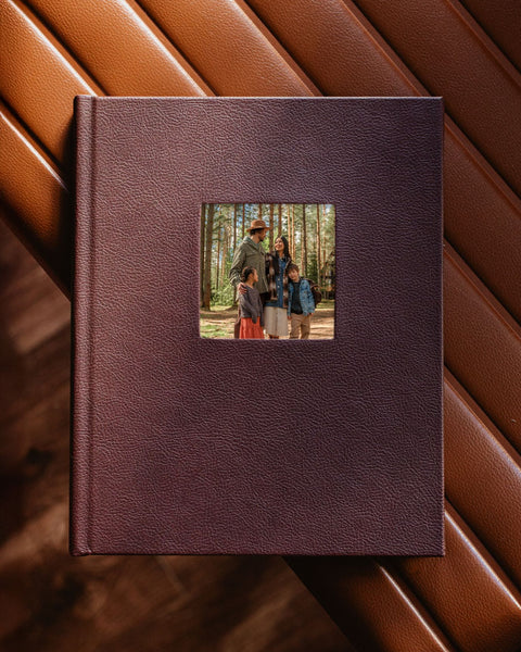 A hardcover photo album with a textured dark plum-brown leather cover, featuring a square cutout on the front displaying a family portrait. The album is photographed from above on a diagonal brown leather surface.