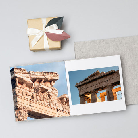 An open photo album lies on a light gray surface, showing two architectural photographs of ancient stone structures under blue sky. A folded light gray envelope is behind the booklet, and a small wrapped gift box with a white ribbon and pastel paper tabs sits above it.